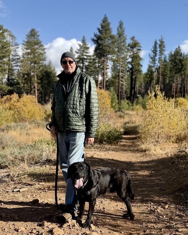 Kevin in a green jacket and knit cap on a dirt trail through pines, holding the leash of his black Labrador, Kukui