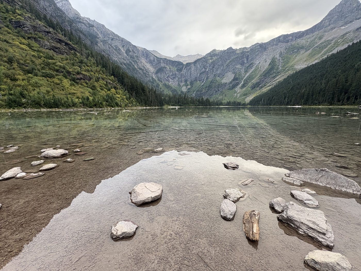 A still alpine lake below grey cliffs, with stones in the foreground