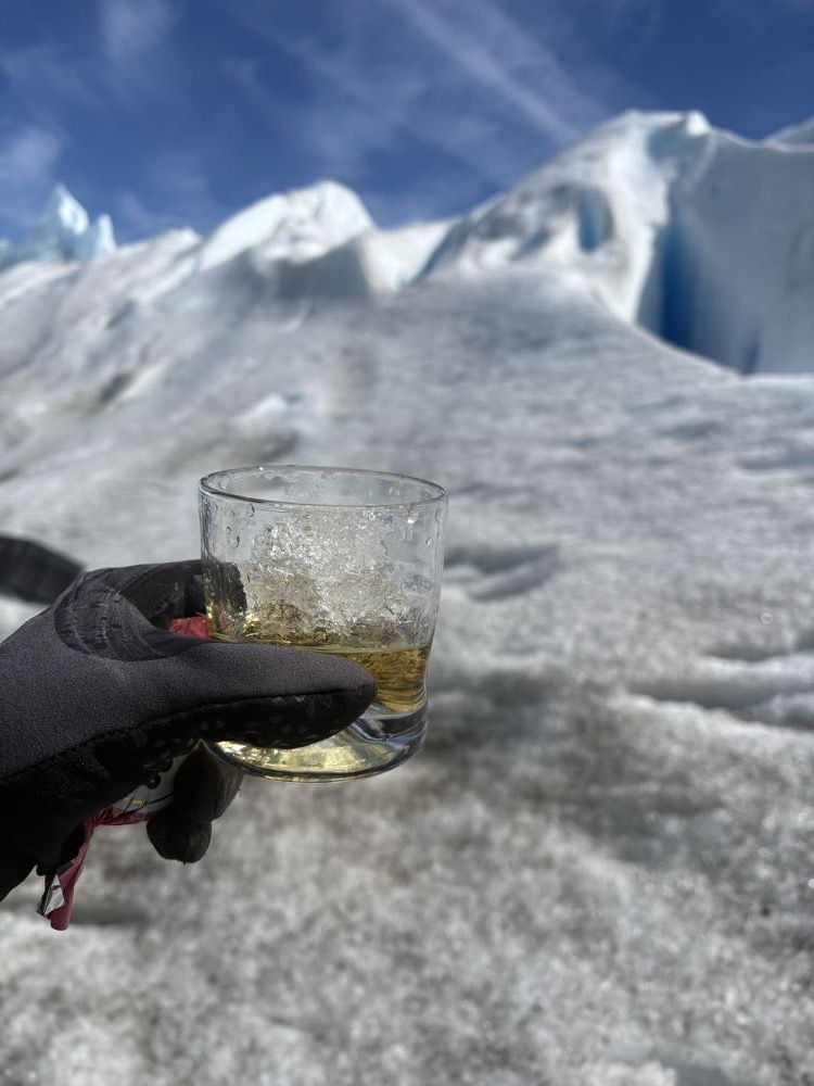 A gloved hand holding a glass of whisky over glacier ice